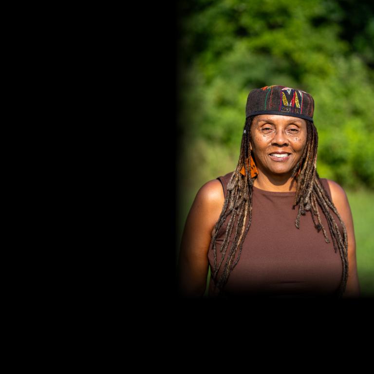 Outdoor portrait of a smiling woman with long gray-streaked locs, wearing a patterned kufi-style cap, orange patterned earrings, and a brown sleeveless top, with a soft green foliage background. Visibilité masquée.