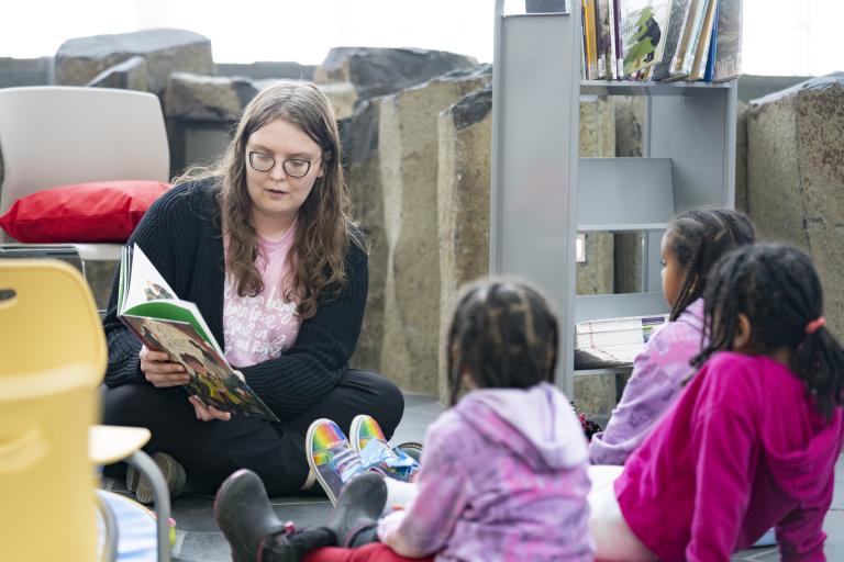 Une personne adulte assise par terre lit un livre à trois enfants assis devant elle. Visibilité masquée.
