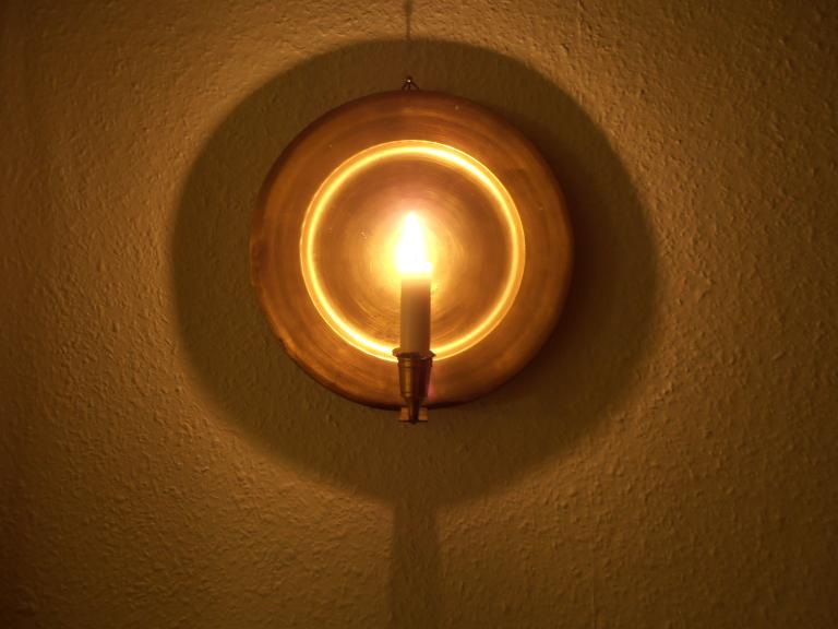 La lueur d’une chandelle sur une applique murale ronde en laiton; son réflecteur brille d’un éclat doré et projette une ombre circulaire sur un mur texturé. Visibilité masquée.