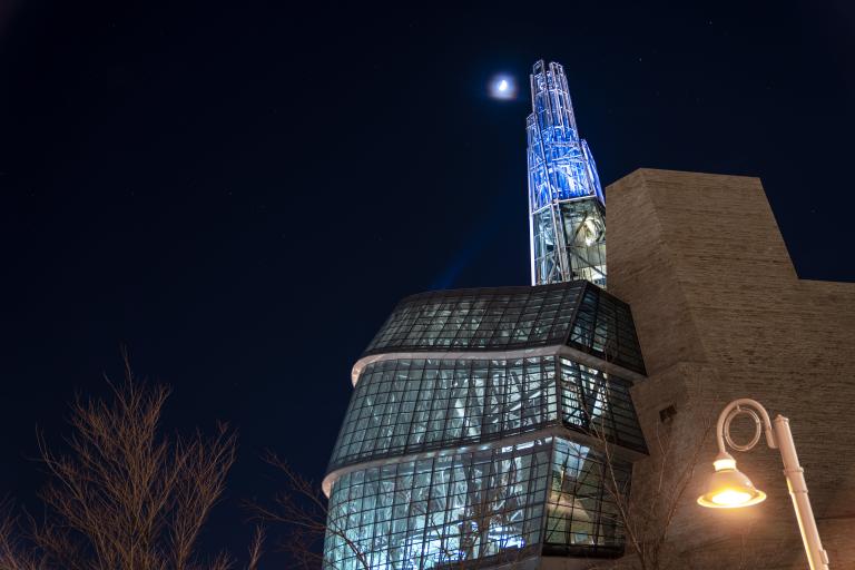 Le Musée canadien pour les droits de la personne la nuit, sa Tour de l’espoir éclairée de bleu se dressant à côté d’un croissant de lune dans un ciel étoilé. Visibilité masquée.
