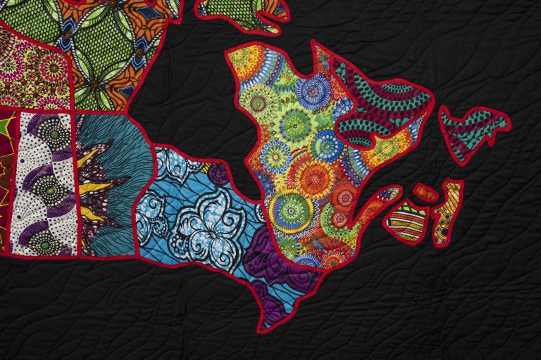 Détail d’une courtepointe représentant une carte de l’est du Canada, chaque région étant découpée dans un tissu aux motifs colorés et délimitée en rouge sur fond noir. Visibilité masquée.