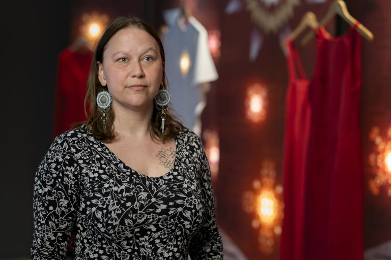 Une femme portant des boucles d’oreilles en perles et un haut à motifs floraux se tient devant des robes rouges suspendues devant un fond rouge foncé, baigné d’une lumière tamisée. Visibilité masquée.