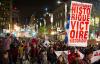 Une mer de jeunes adultes marche dans une rue animée le soir, entourée par des bâtiments et des lampadaires. Une personne tient une pancarte où l’on peut lire « mouvement historique victoire historique ». Une autre porte un costume de panda.