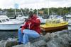 Un homme portant un survêtement de couleurs vives et une calotte blanche est assis sur des rochers, avec des bateaux et un port en arrière-plan.